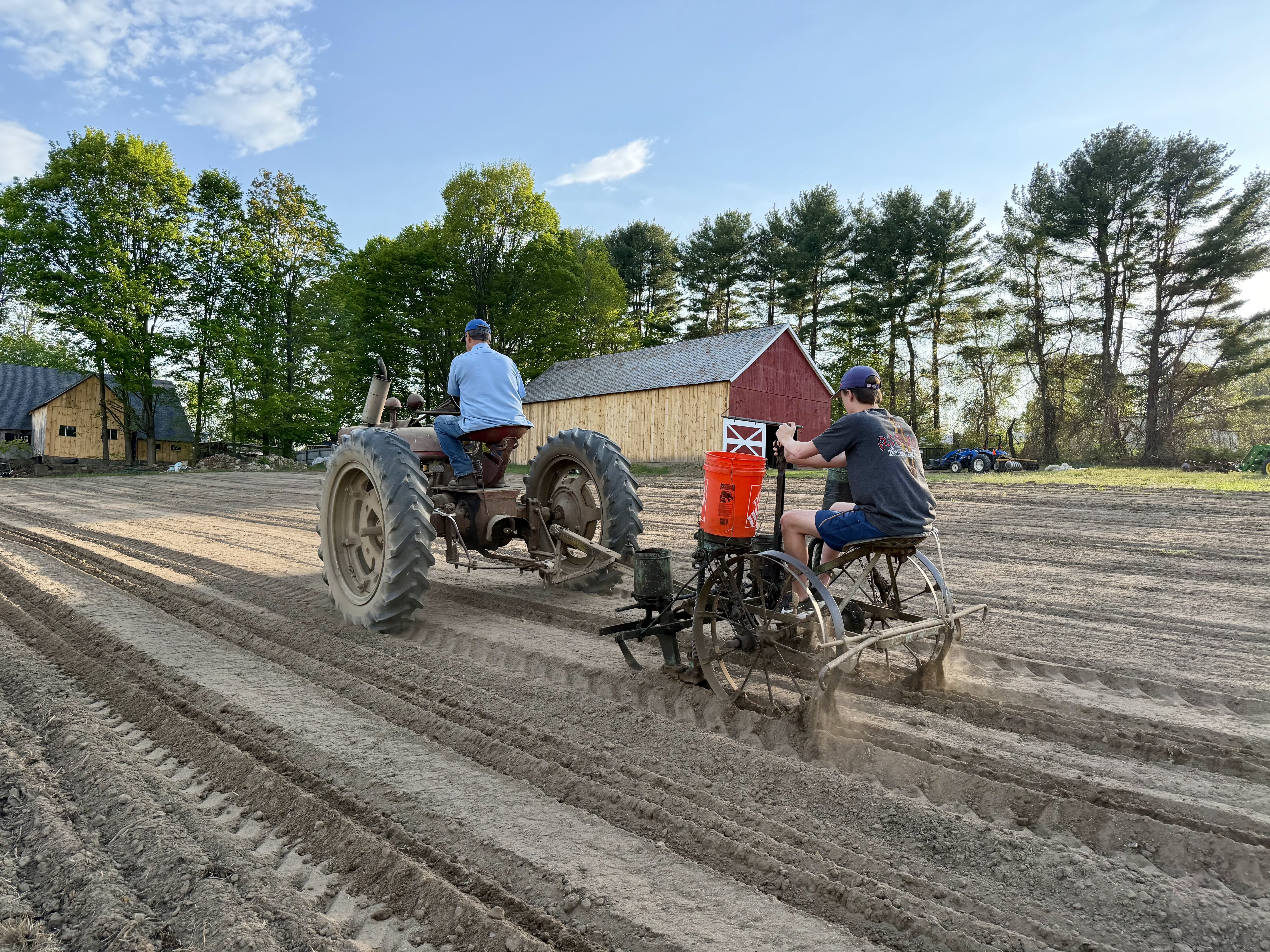 Farm workers planting corn with a tractor in a freshly tilled field beside red barns.